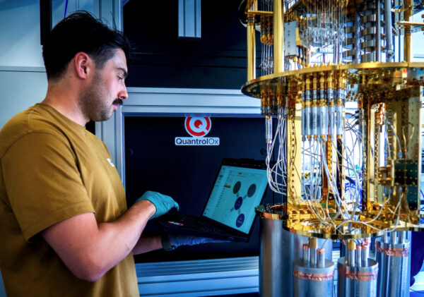 QLX › QBN - Quantum Business Network › QBN - Quantum Business Network Technician in a brown shirt using a laptop to monitor a gold-colored laboratory instrument labeled QuantrolOx.