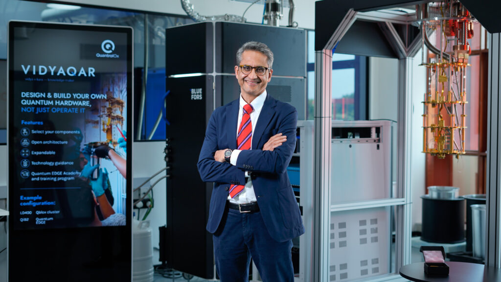 Vishal-03 › QBN - Quantum Business Network Smiling businessman in a navy suit with a red tie stands with arms crossed in a high-tech lab behind him.